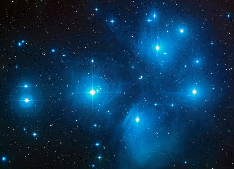 A close telescopic photo of seven stars in the foreground with a dense cluster of others surrounding them, with more behind.