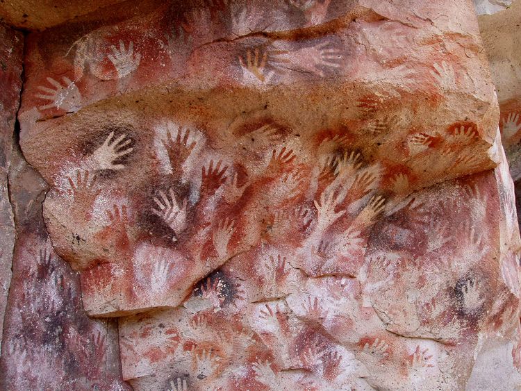 The silhouettes of hands are traced on a rock wall, using various red, brown, and white paints.