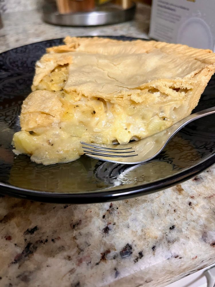 A slice of pie with moist, golden filling speckled by black pepper. It sits on a black ceramic plate with a fork nearby.