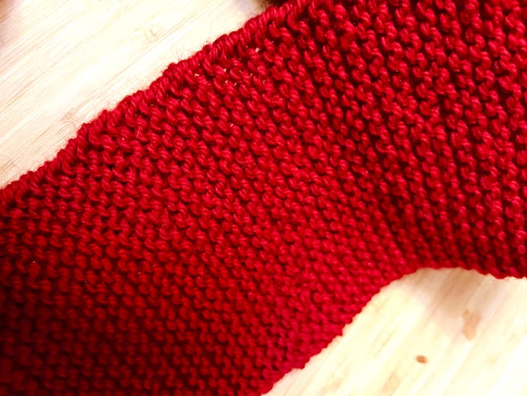 A section of a red wool scarf, shown in closeup over a wooden surface. The scarf is knit with garter stitch.