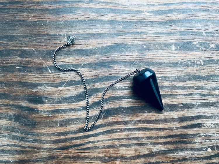 A black conical pendulum on a metal chain capped with a clear bead. It all lies on a wooden surface.