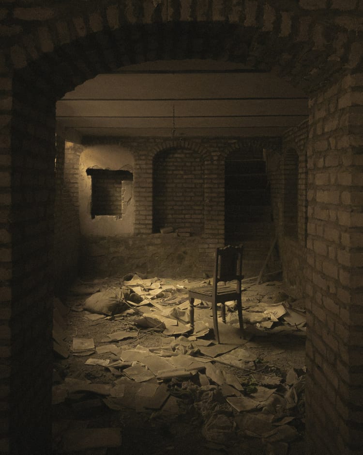 A sepia tone photo of what looks like an abandoned brick basement, with a vacant chair and detritus strewn everywhere.