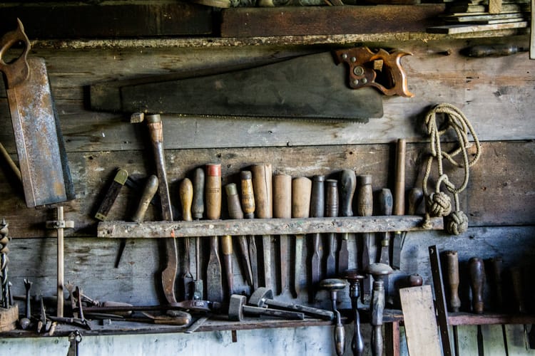 An array of antique tools displayed against a background of wooden planks. The tools include a saw, chisels, awls, and more.