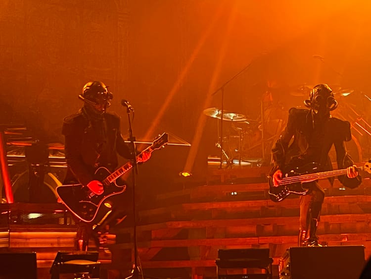 Two people wearing all black attire and stylized gas masks play electric guitars on a stage. Heavy orange lighting.