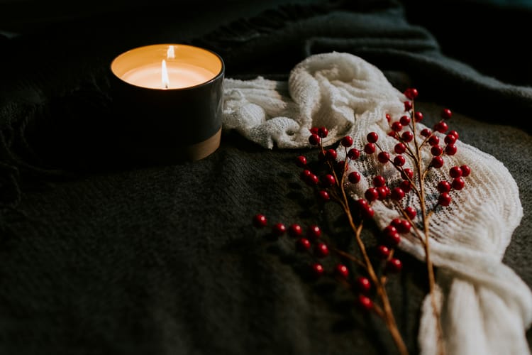 A small, lit candle in a dark container sits on some dark fabric near a white scarf and two sprigs of American holly.