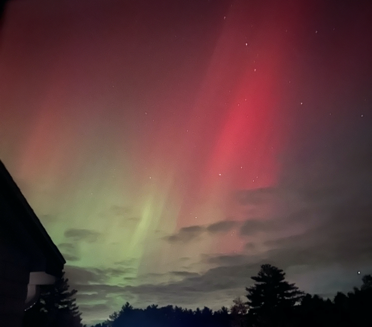 A night sky with stars and clouds, but more prominently enormous bright red and pale green streaks of the Northern Lights.