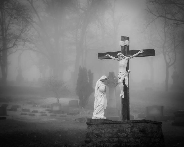 Black & white photo of a cemetery tomb with Jesus crucified while his mother Mary looks on. The rest of the graveyard is very foggy.