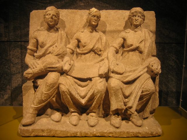 A terracotta relief of three female-coded figures who are seated with various objects in their hands.