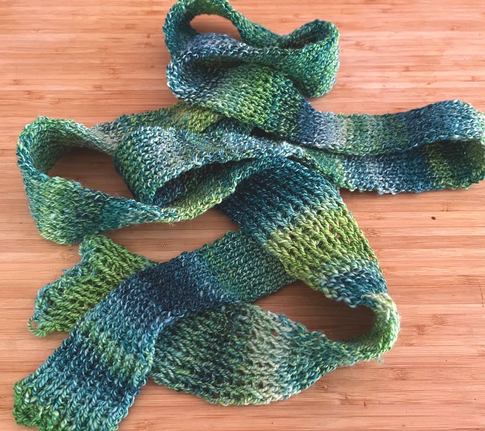 A scarf colored in irregular bands of numerous green and blue shades. It's very long. A rib stitch has been used.