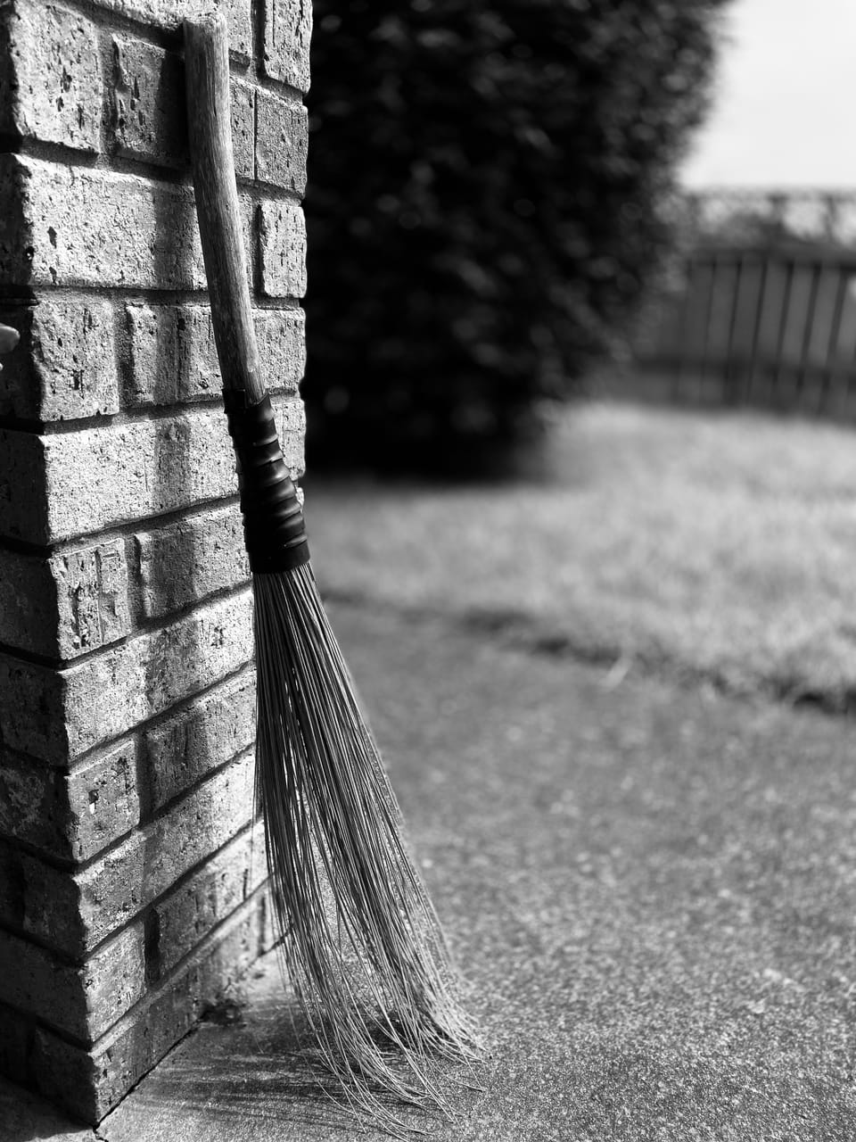 A black and white image of a reed broom bound to a wooden handle. It leans against a brick wall outdoors.