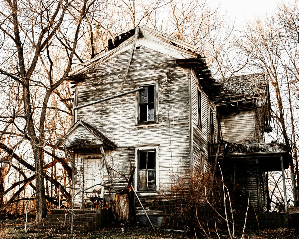 A decrepit New England-style colonial house with decaying white siding, surrounded by bare trees.