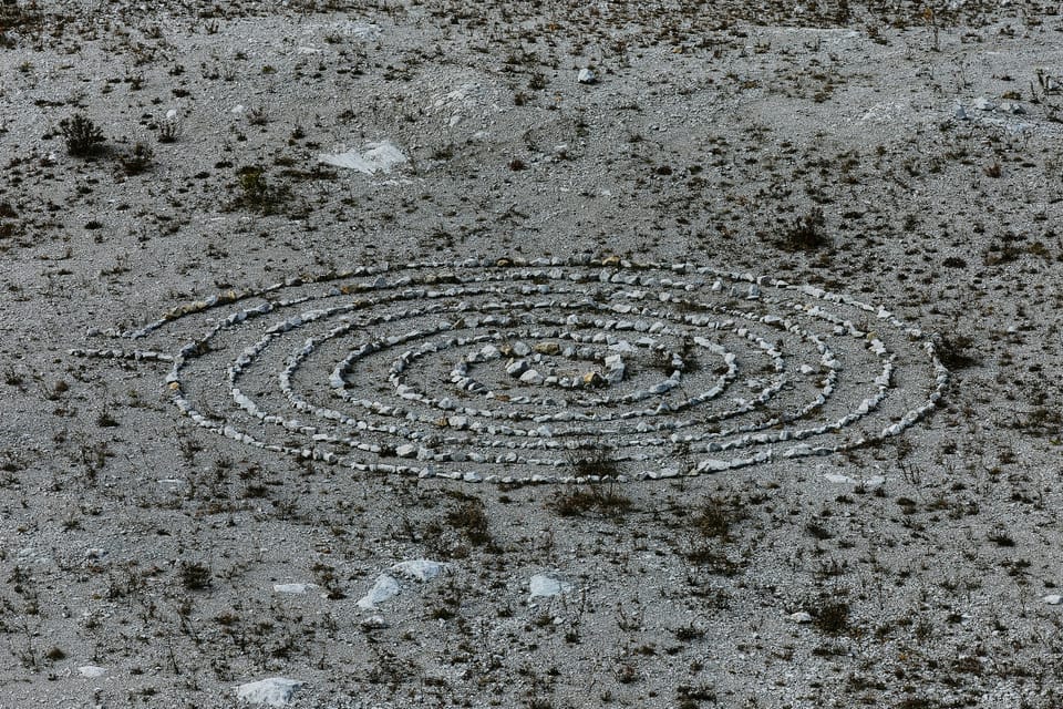 A spiral of stones created on grey dirt spotted by scrubby bushes.