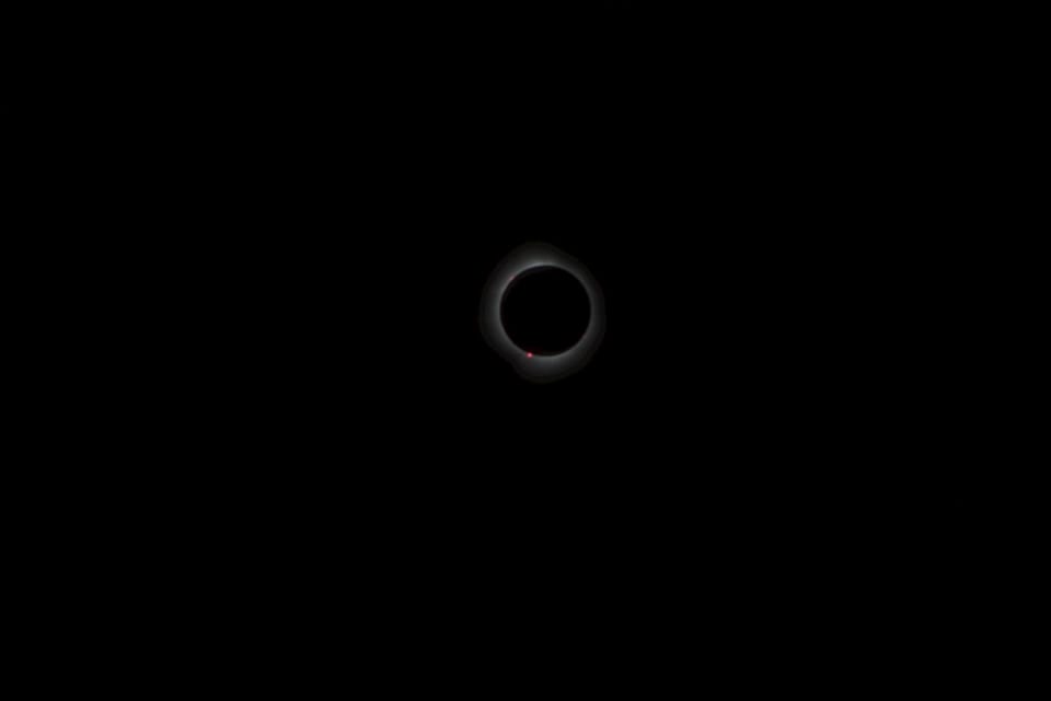 The Sun's ghostly white corona around the black disc of the Moon with a red gem at bottom. Black sky due to camera limits.