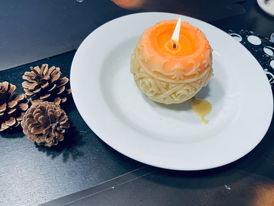 A lit, round beeswax candle carved with floral-ish motifs. It sits on a white plate with a few decorative pinecones nearby.