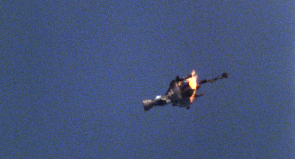 A blurry film frame of a burning rocket engine suspended in blue sky.