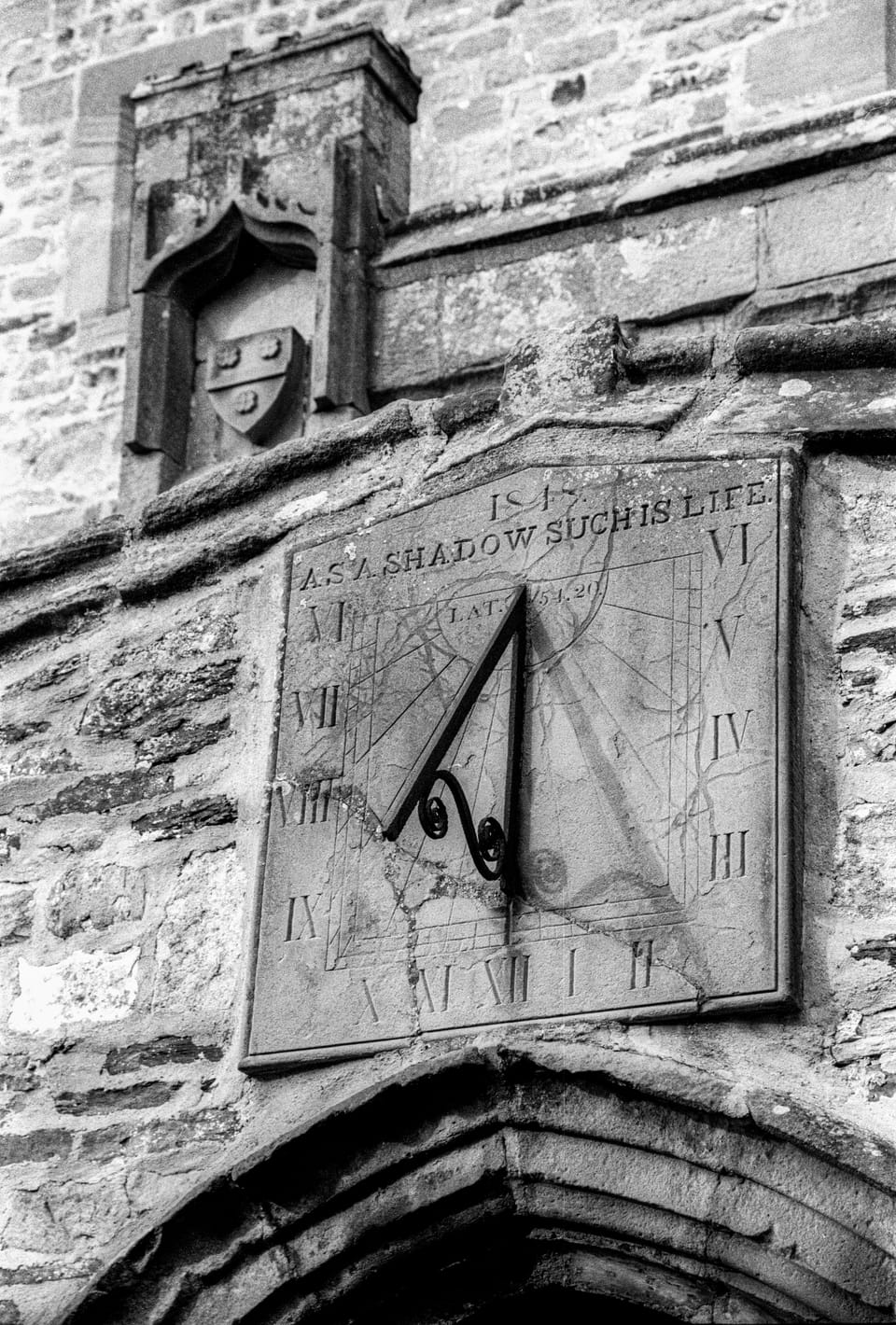 Black and white photo of a wall-mounted sundial, hours marked with Roman numerals. Top text reads "as a shadow such is life."