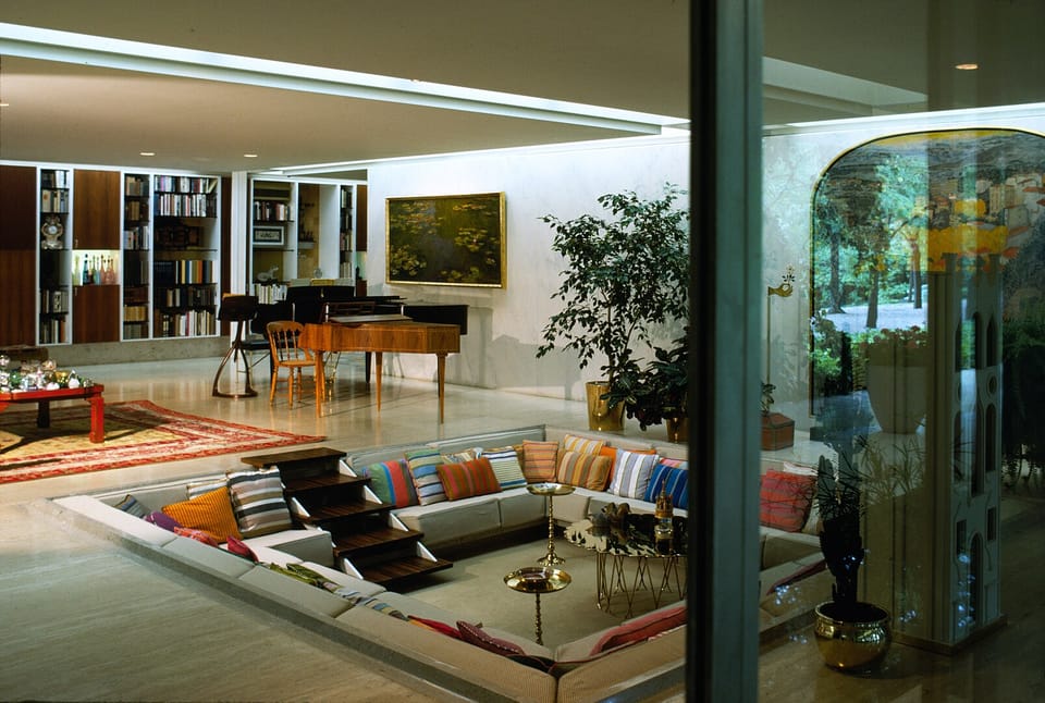 A spacious, open room with white walls, pale wooden floors and tasteful furniture that gives way to a square seating pit with colorful cushions.