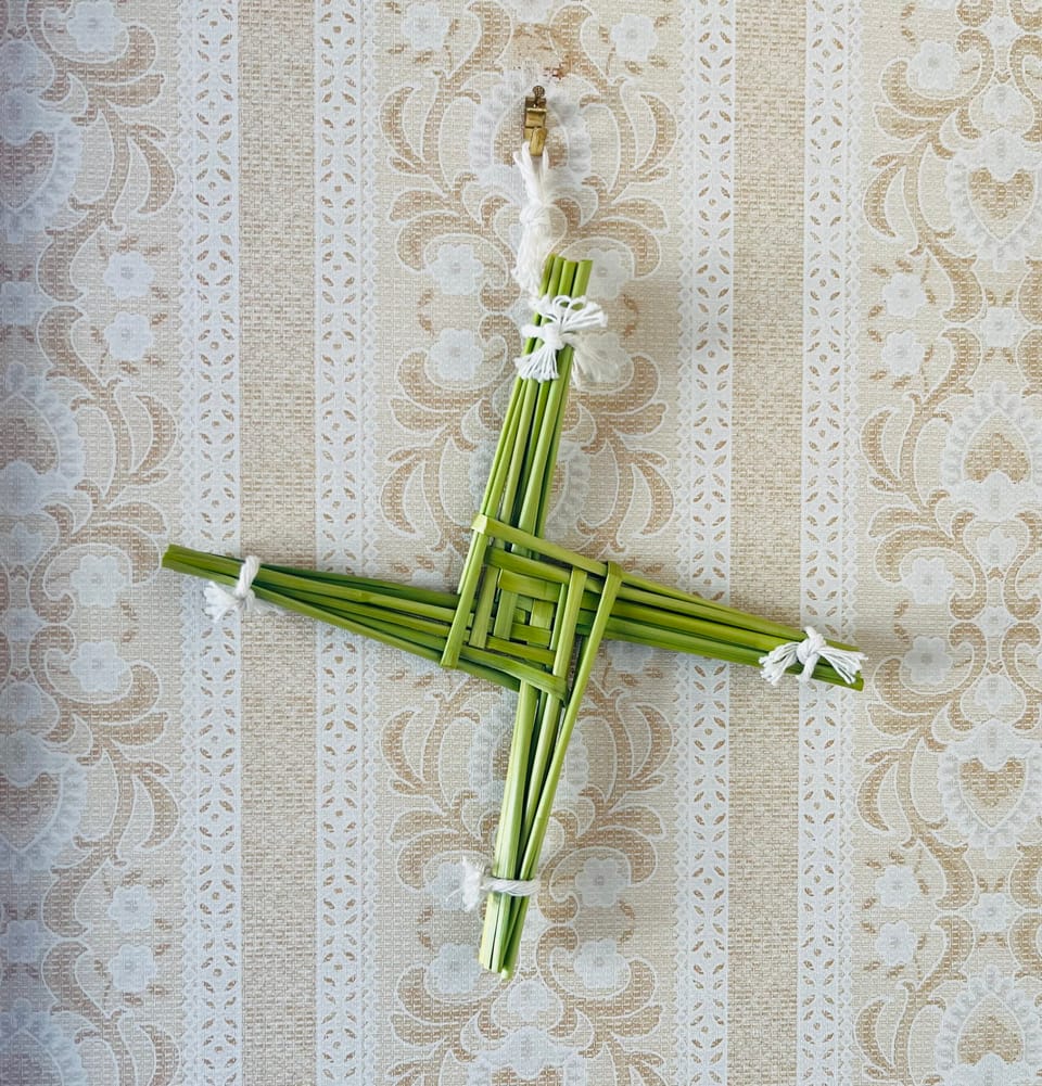 A piece of straw art. It looks like an equilateral cross where the arms seem to emerge from a squared spiral formation in the middle. The straw is green.
