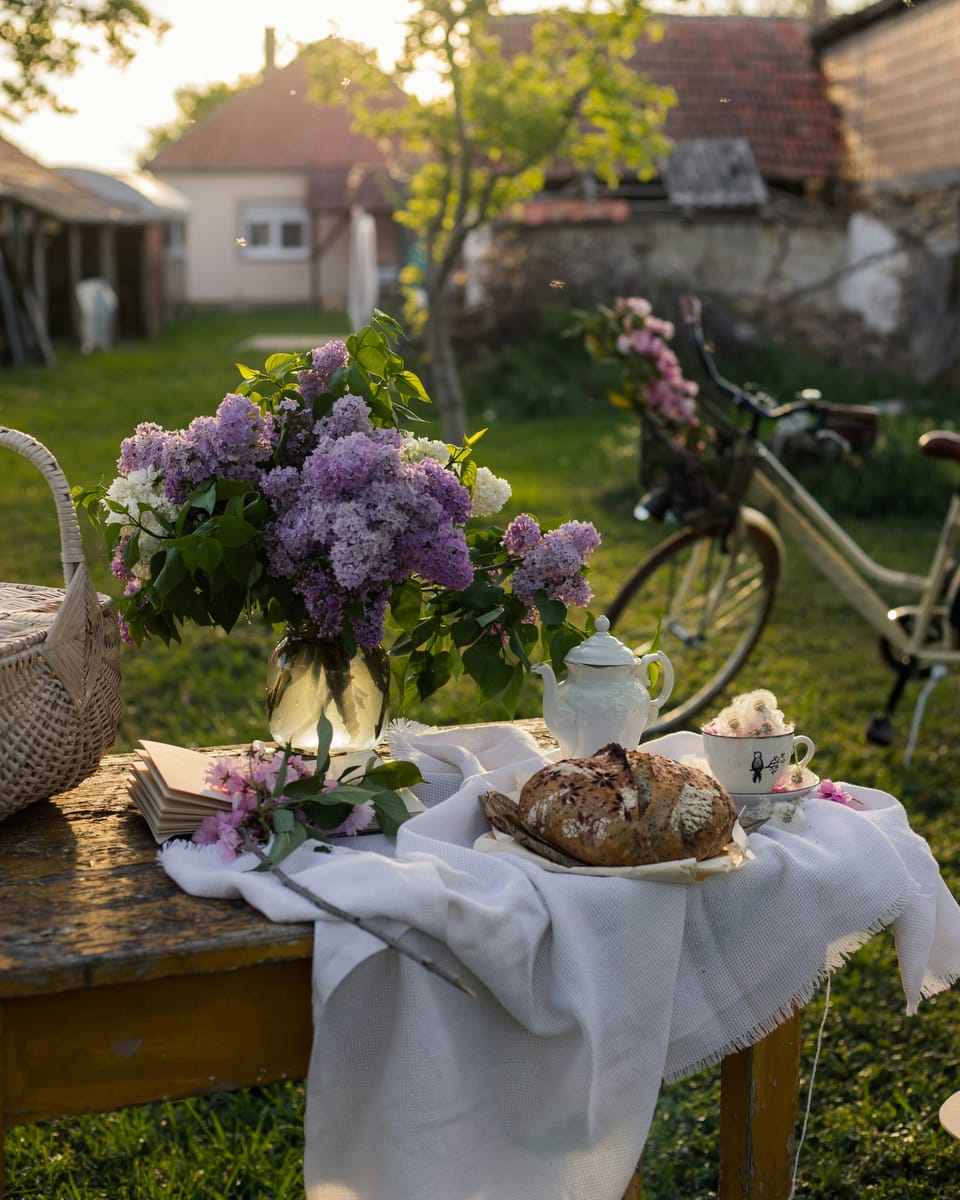 A twee photograph shot at the golden hour. It shows bread, a tea cup and pot, an open book, and some purple flowers on a white cloth, among other items.